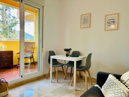 Sala de estar con mesa blanca, sillas grises y acceso a un balcón iluminado.